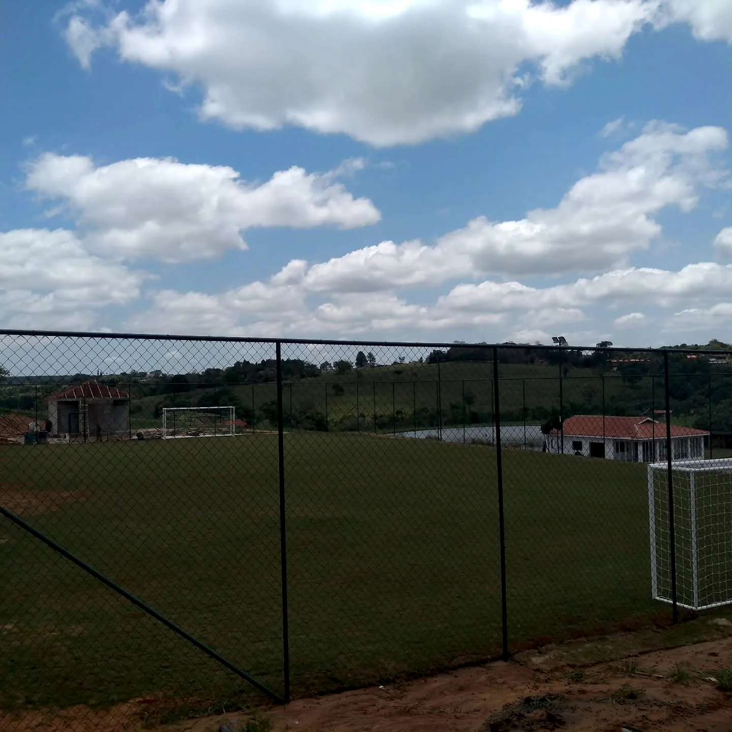 Campo gramado visto através de alambrado, com traves e área esportiva ao ar livre.