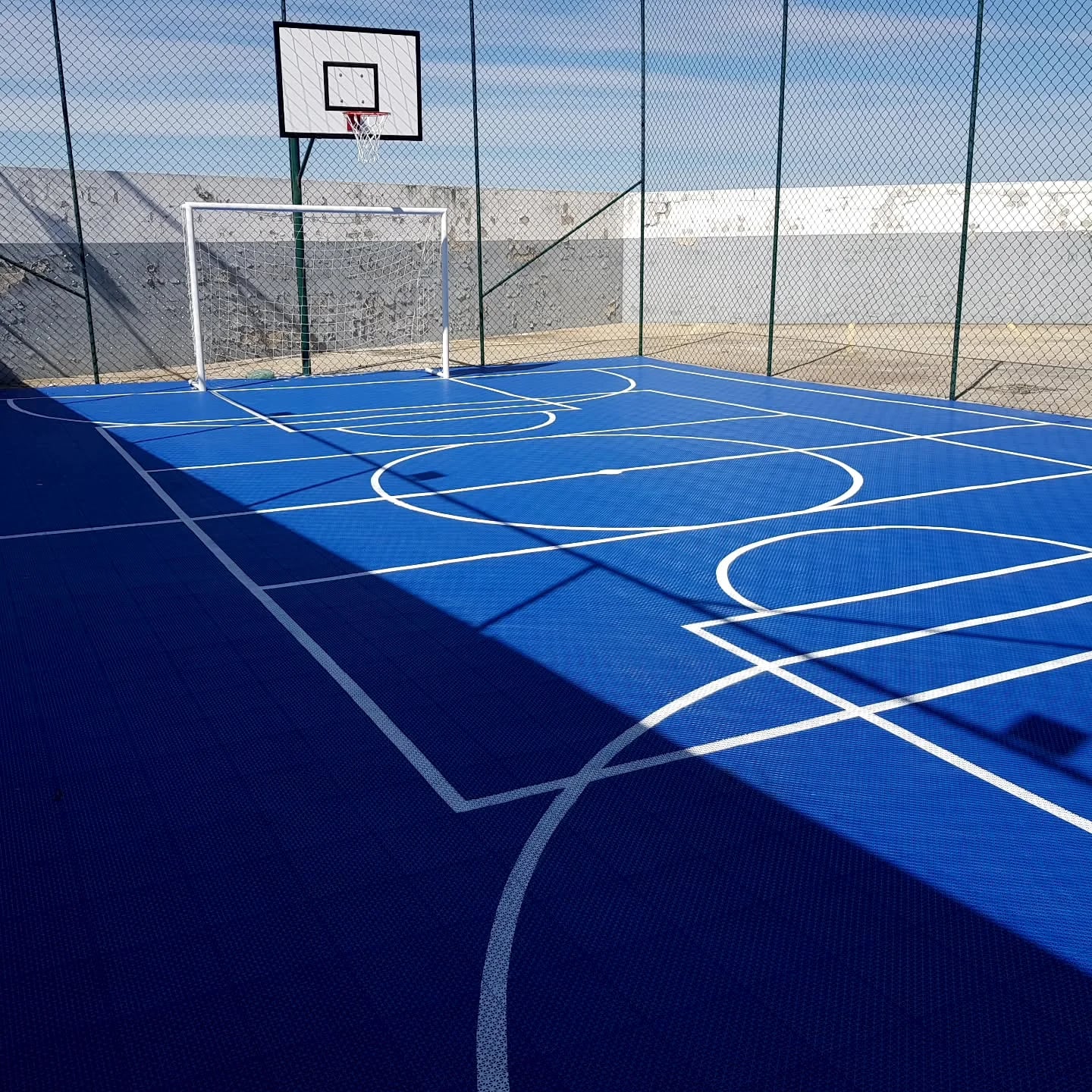 Quadra externa azul com marcações esportivas, gol, tabela de basquete e alambrado.
